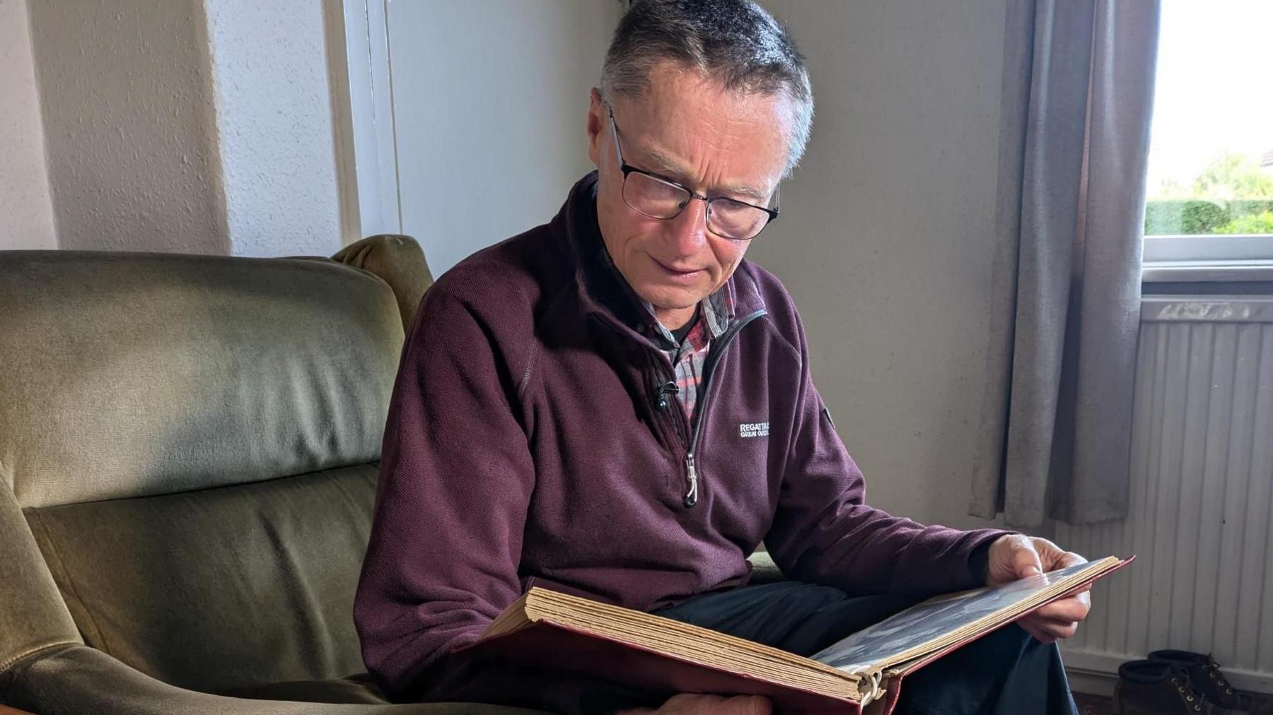 Andrew, a man with short grey hair and glasses looks through a family photo album. He is wearing a burgundy zipped jumper and sitting on a brown armchair in his living room.