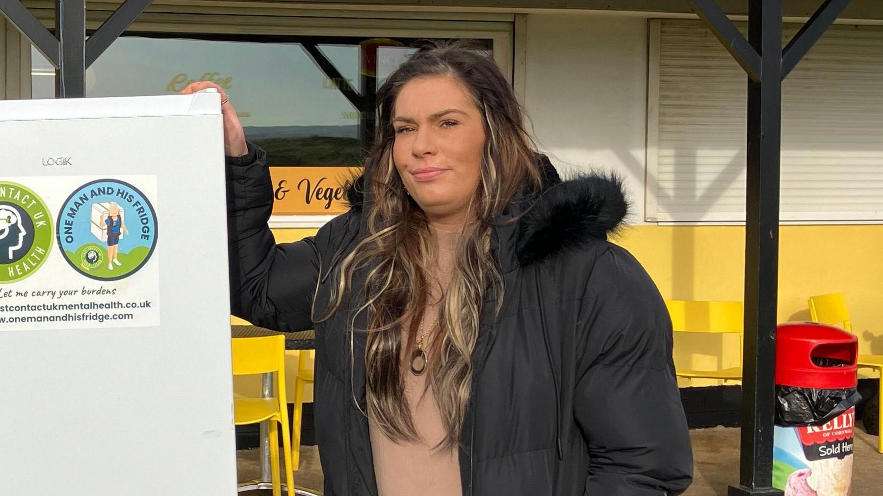 Stacey Baldam who long wavy, blonde, black and brown hair is standing next to a fridge - which has stickers on it saying One man and his fridge.