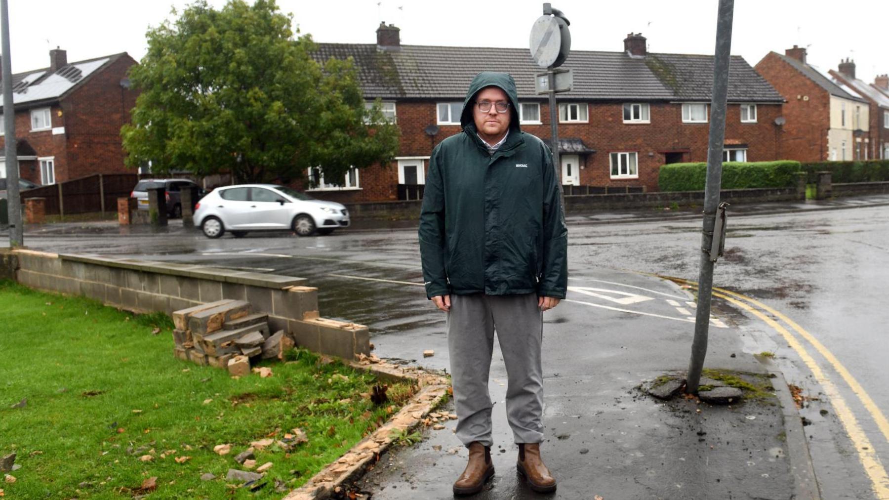 Eric Maw stands in front of a bungalow next to the remains of a garden wall with rubble and bricks on a lawn and a bent grey-coloured signpost

