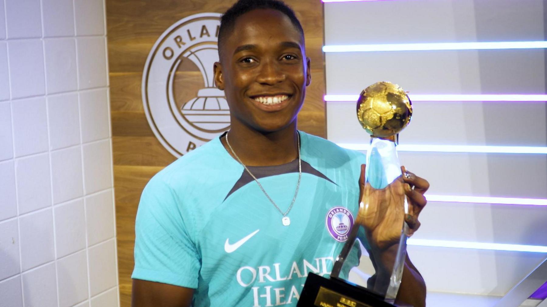 Barbra Banda, wearing an Orlando Pride training top, smiles as she holds the BBC Women's Footballer of the Year award, with her club's logo on the wall behind her