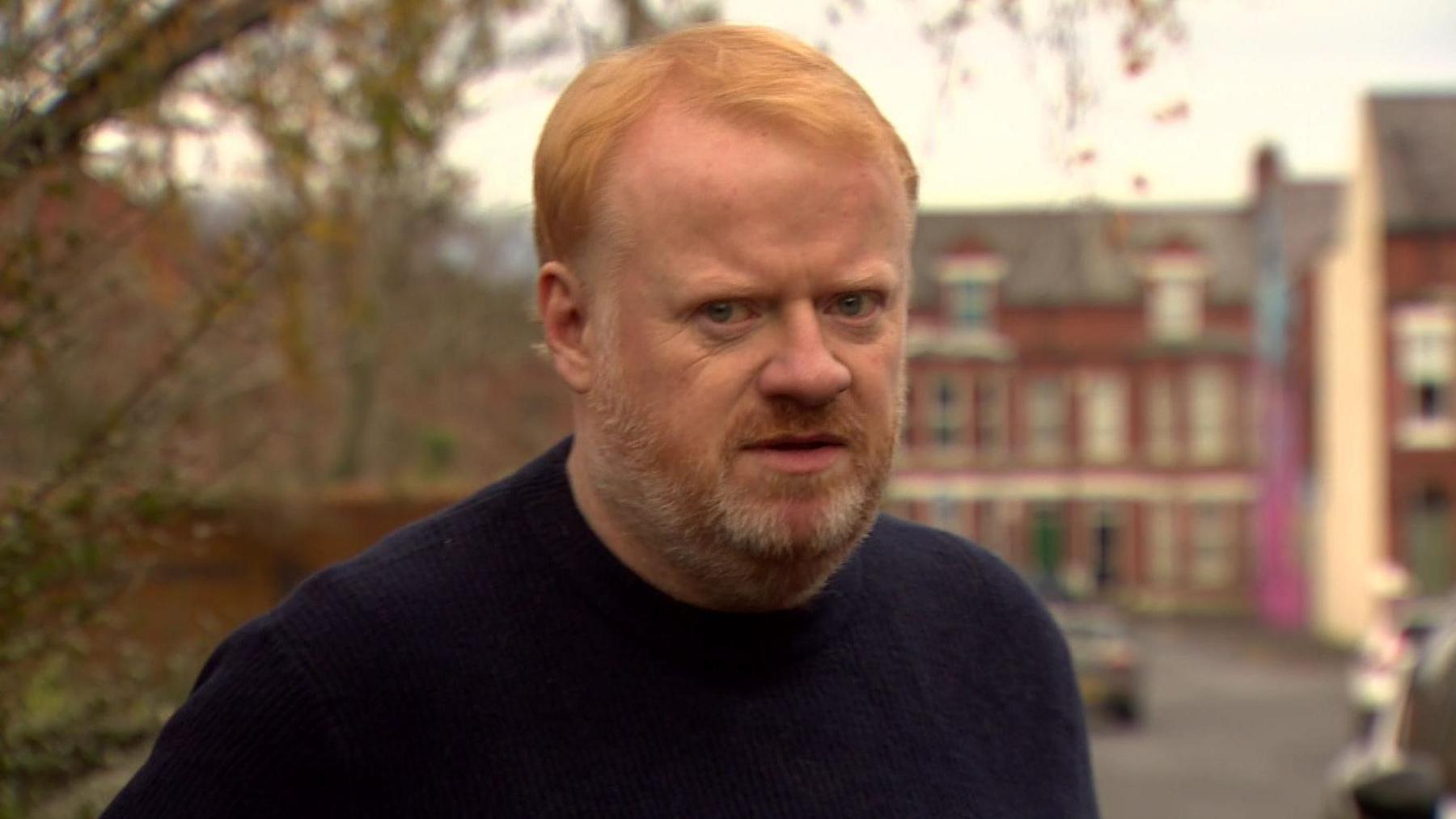 A red haired man with a red and white beard is wearing a navy top and looking to the camera. Behind him out of focus is a street with red brick houses and cars. A tree can also be seen behind him. 