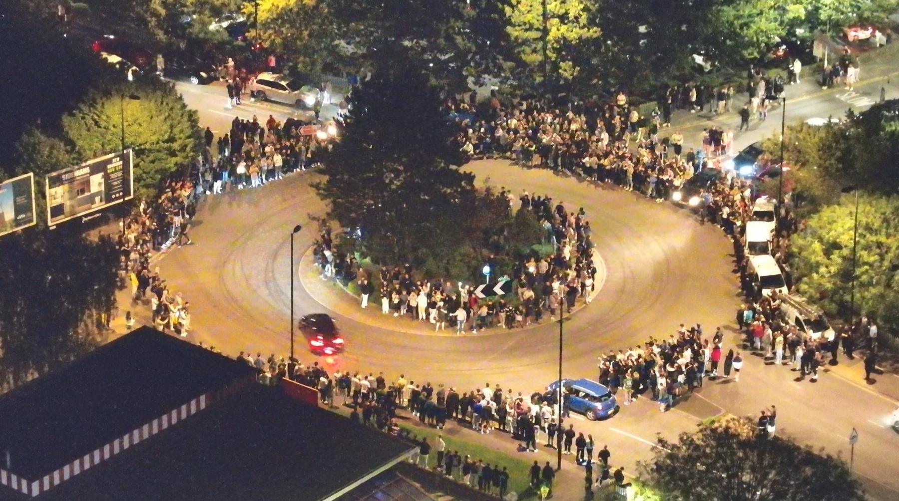 An overhead shot at night time of cars going around a roundabout whilst crowds line the pavement.