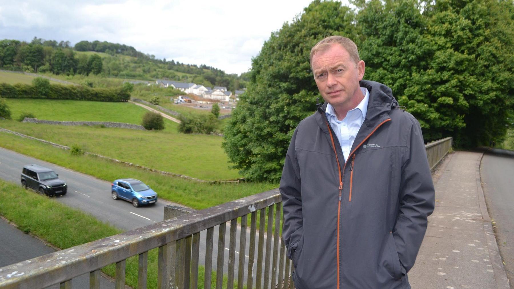 Tim Farron standing by the bridge on Brigsteer Road. He has short fair hair and is wearing a light blue shirt and grey jacket. Below him, two vehicles are being driven along a road. In the background are fields and trees.