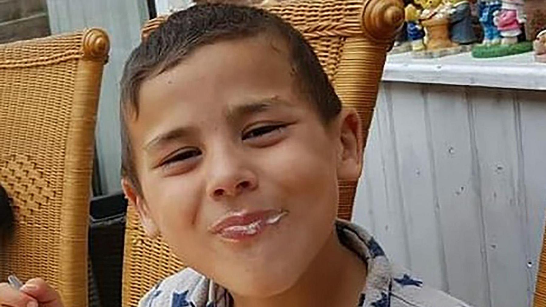 A boy sits on a wicker chair outside and smiles at the camera. He has dak brown hair and he is eating ice cream.