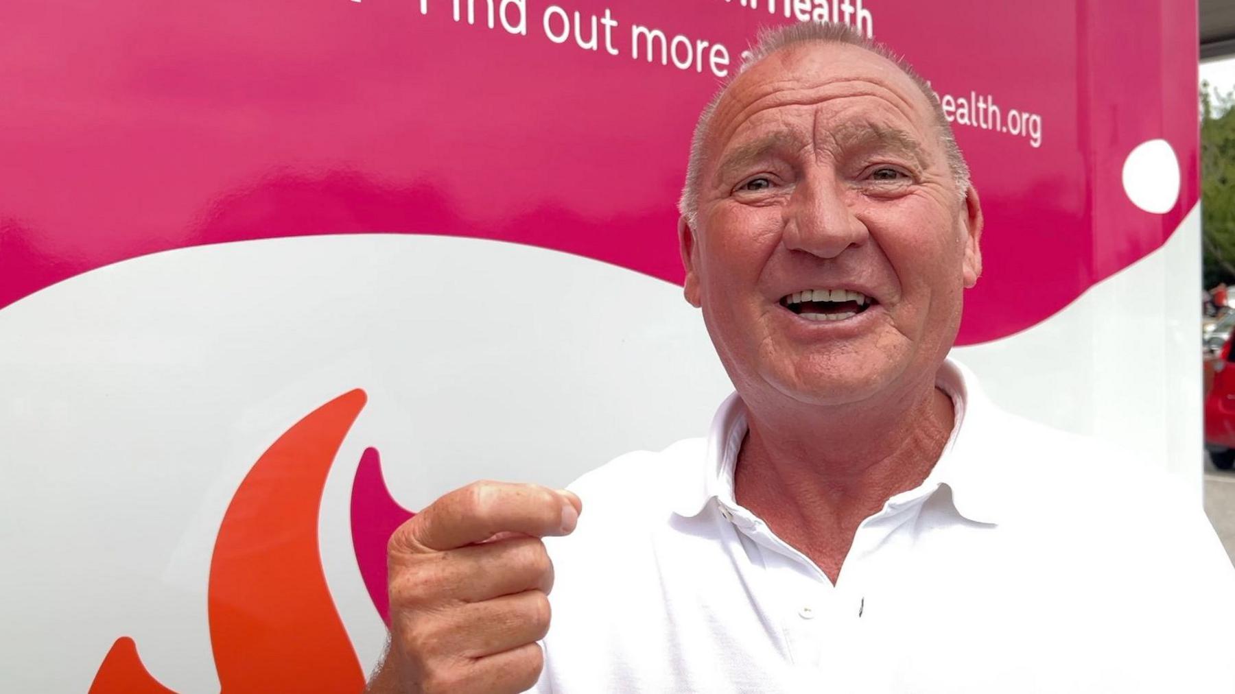 John, wearing a white shirt, standing in front of a pink and white clinical testing van.
