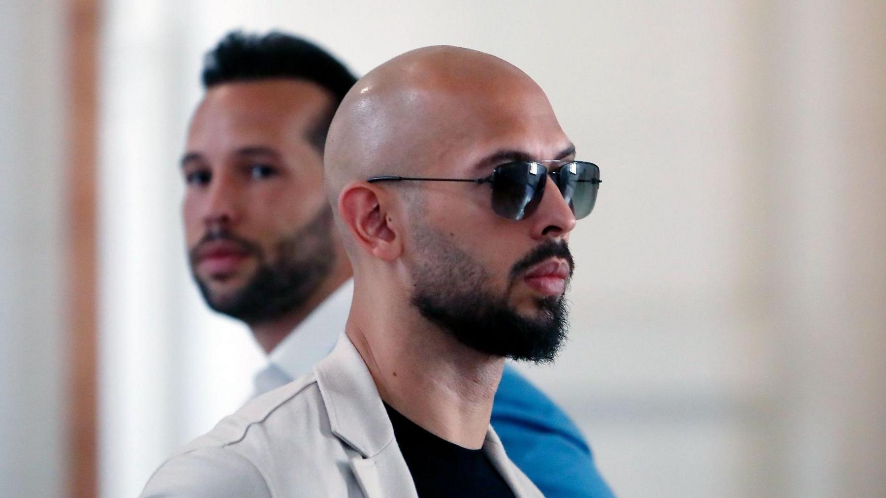 Andrew Tate facing to the right, wearing aviator style sunglasses, his head is shaved and he has a dark moustache and beard and wears a black T-shirt and white jacket. His brother is standing next to him facing the other way.