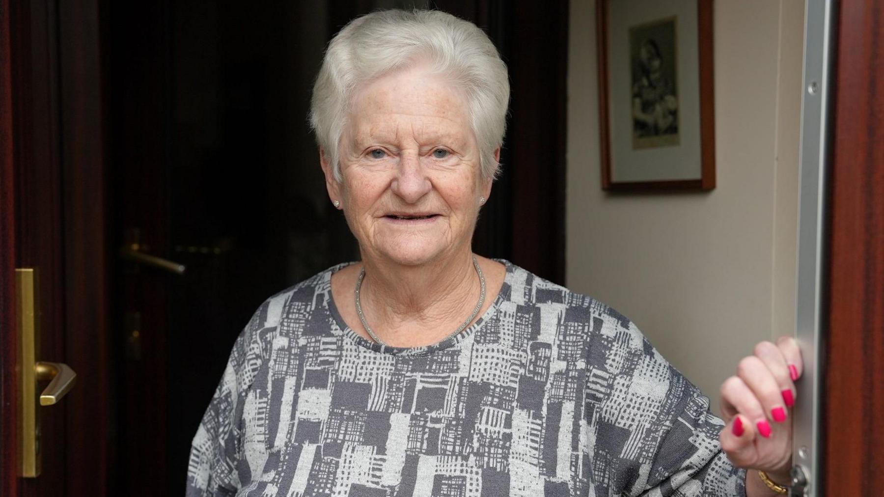 Ruth Pressley standing in a doorway. She has short smartly coiffed white/grey hair, and pink fingernails. She is wearing a grey necklace and grey chequered top. There is a framed photo on the wall behind her and the doors are dark brown with brass handles. 