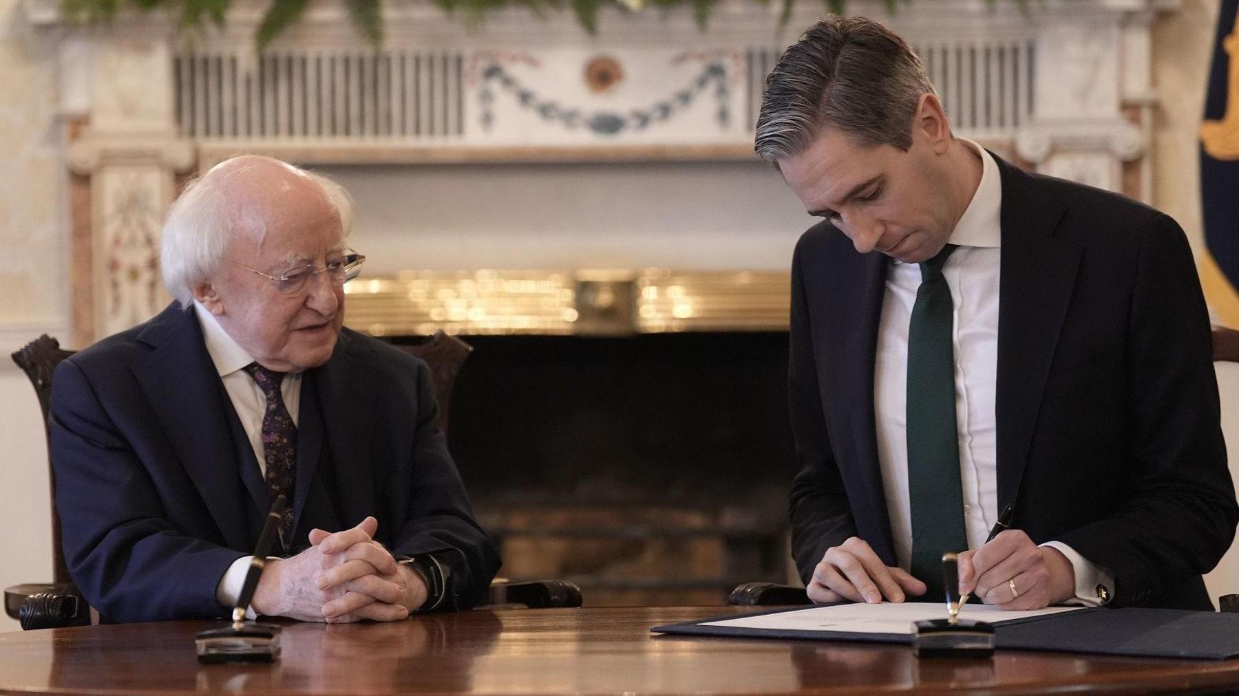 Michael D Higgins - an older man with white hair wears rimless glasses is sat at a desk with Simon Harris. He has short dark hair and is signing a piece of paper. Bother men are wearing dark suit jackets, white shirts and ties.