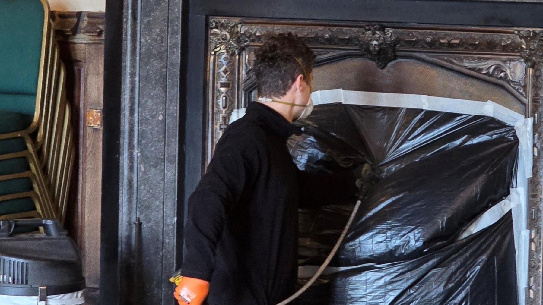 Matthew Taylor with brown hair wearing a black top with his back to the camera cleaning a chimney. He has taped up the black fireplace and wearing a mask and gloves.  