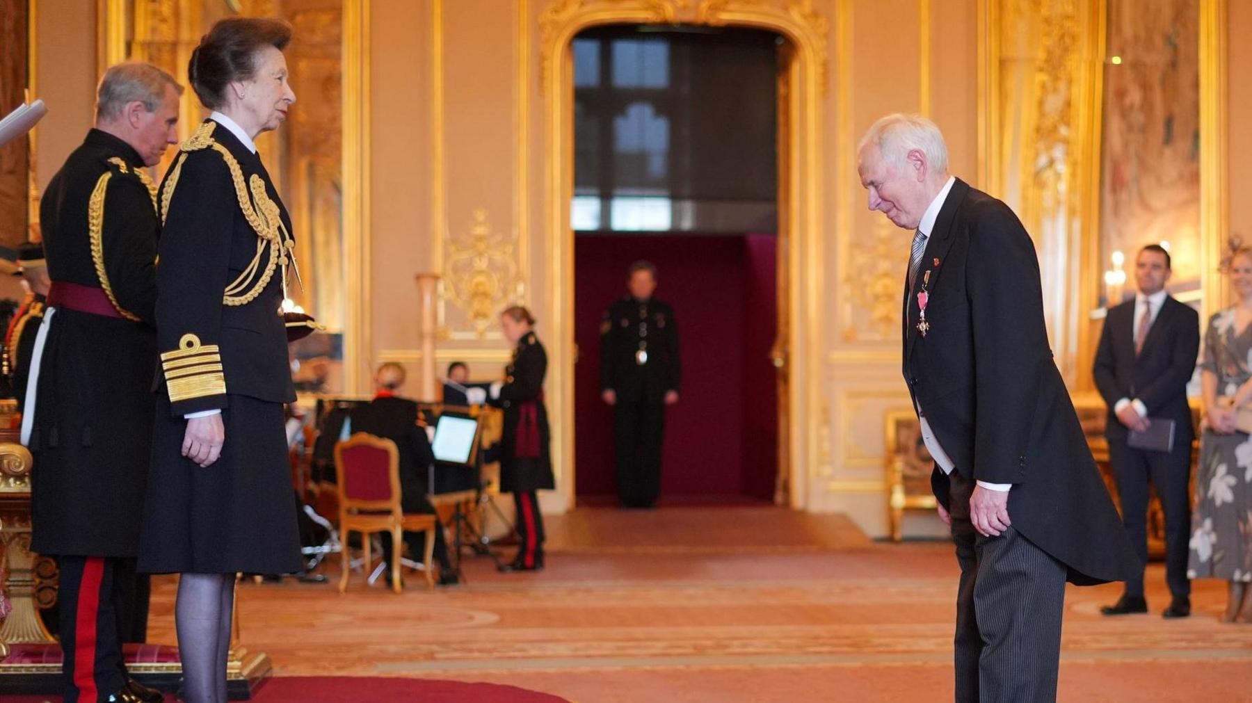 Nick Owen bowing to Princess Anne. The Princess Royal is wearing ceremonial uniform with gold trim and several other people are in the background, also dressed in military uniform. 