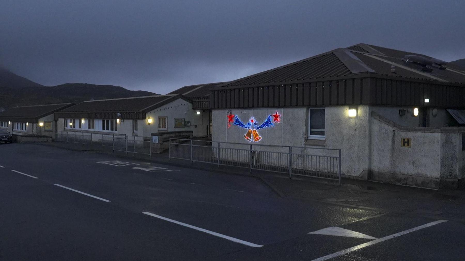 There are Christmas lights lit up on a wall of the school. It is dark and there are some lights illuminated along the walls of the school buildings.