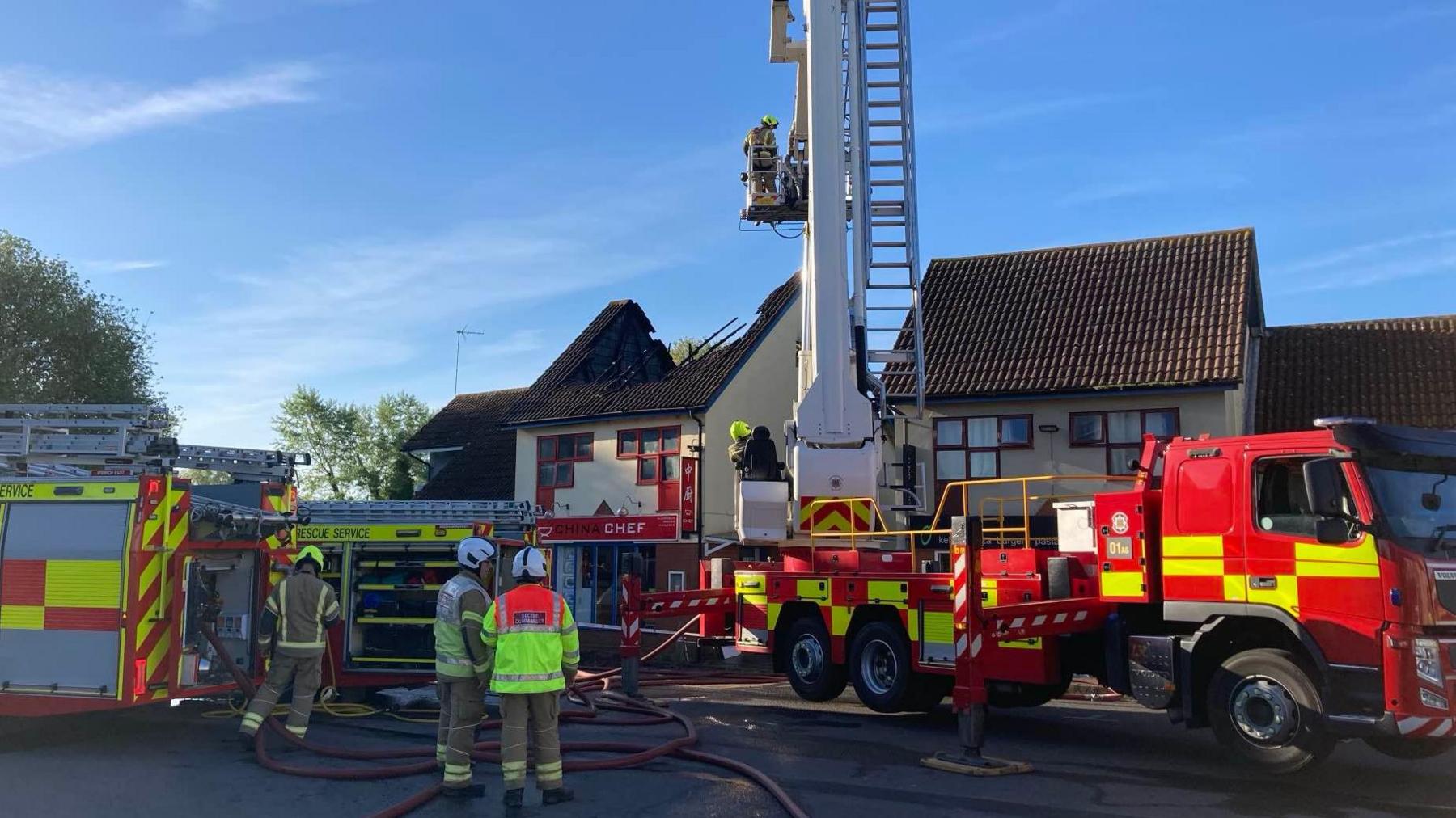 Fire crews outside a property. An aerial ladder can be seen in use. Firefighters watch on. A property can be seen where its roof has been burnt and damaged in a fire.