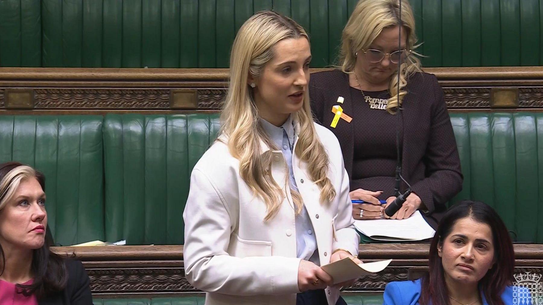 A woman in a white blazer and blue top stands in the House of Commons talking. 