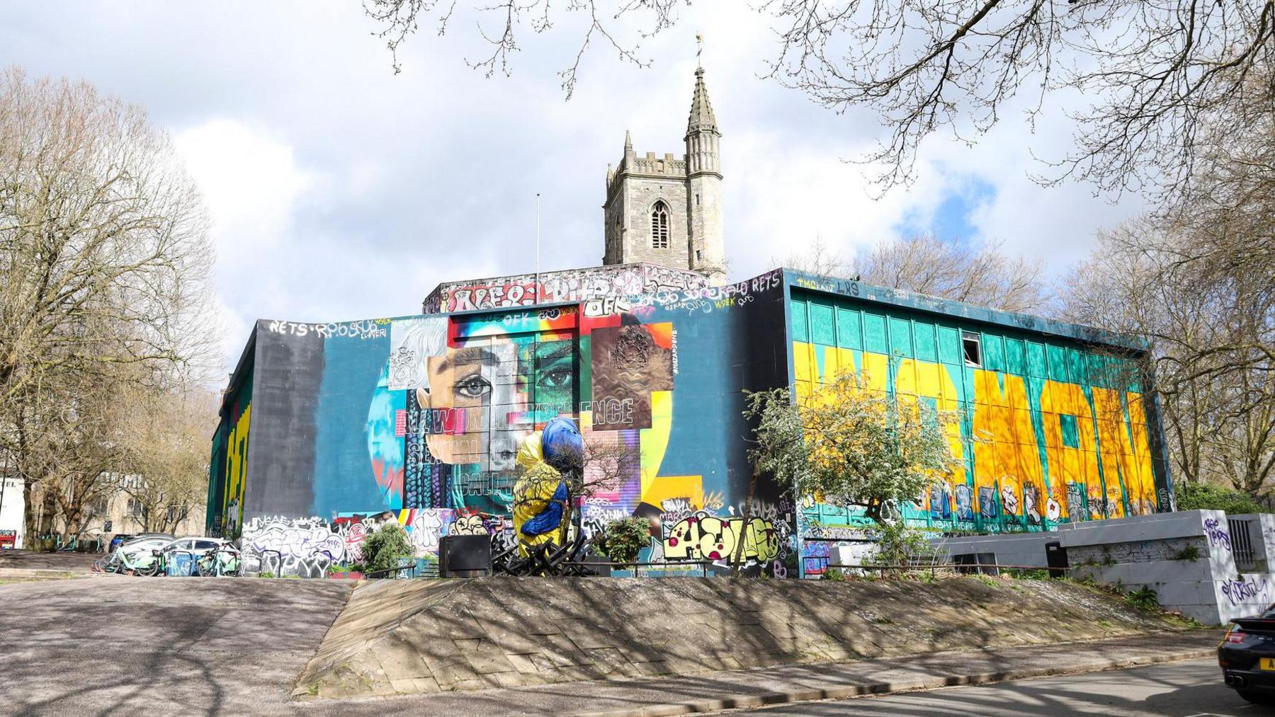 A view of the old Bank, in the centre of Bristol next to Castle Park, covered in bright colourful murals and artwork. On the Right are large yellow words across the building that say "Woman". On the corner is artwork that includes a face.