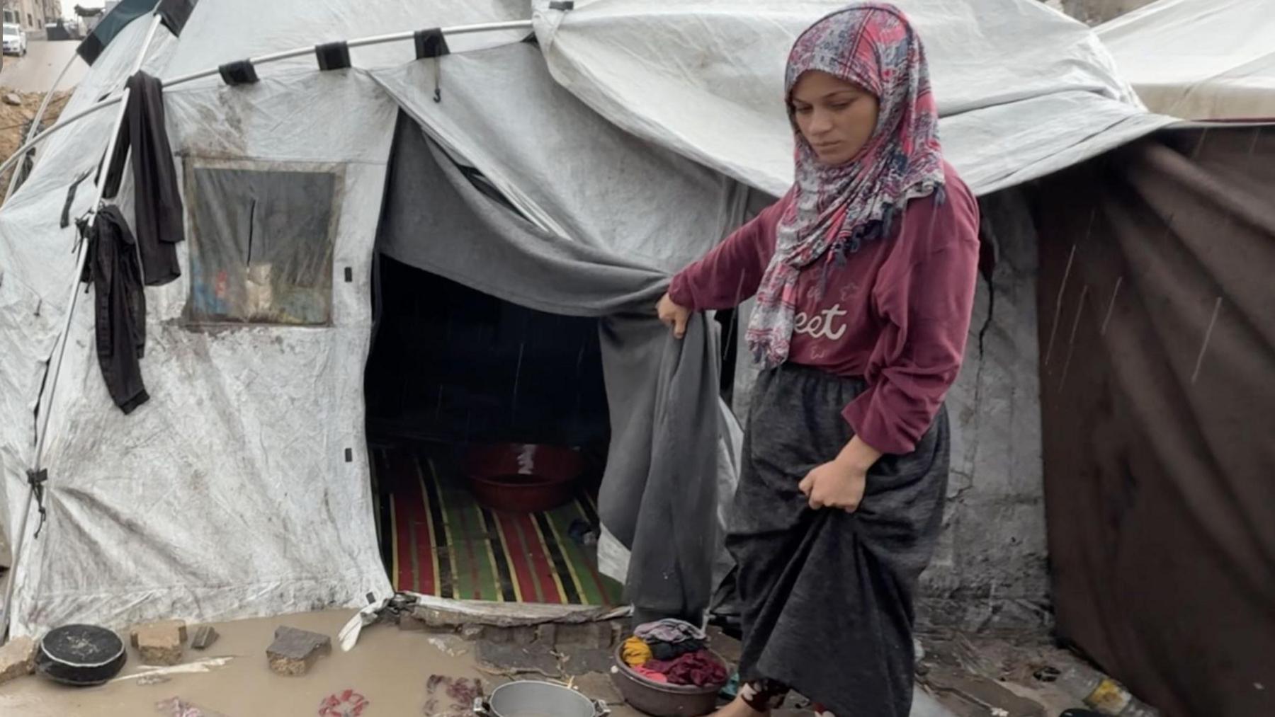 Palestinian woman, standing in wet and mud, holds open the door of her tent 
