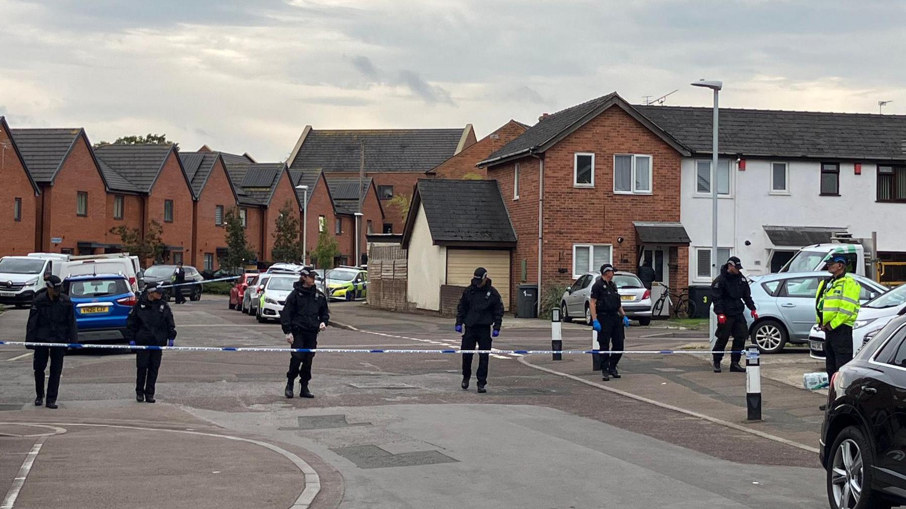 Police officers, dressed in black, conduct a search behind a police cordon on a residential street. 