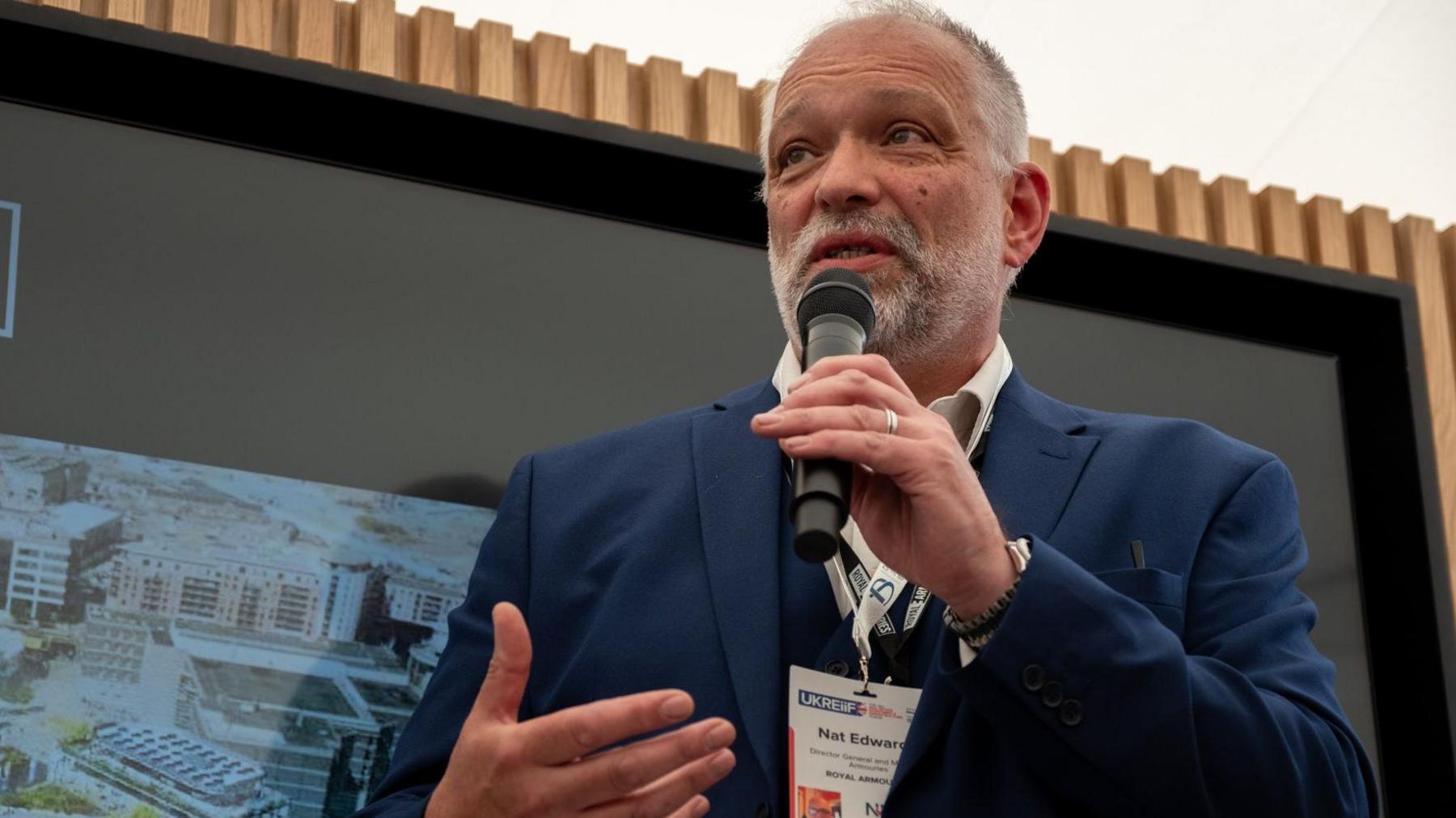 A man with short grey hair and beard wearing a blue site and white shirt with a lanyard around his neck