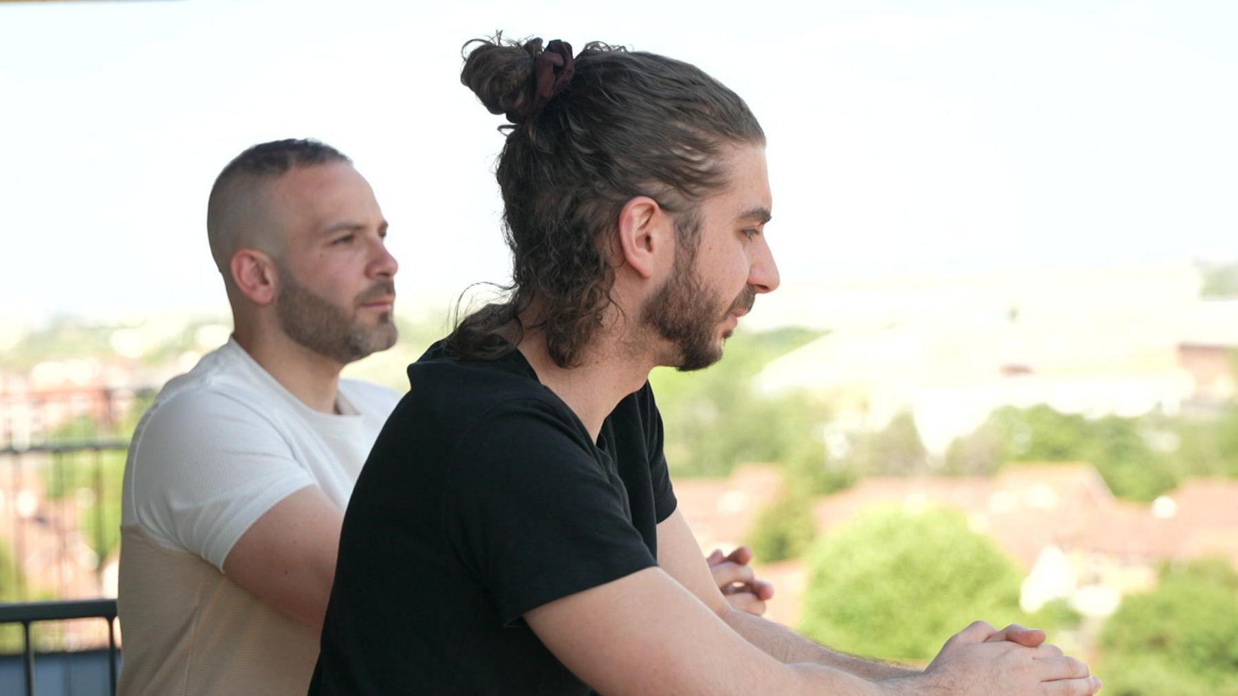Two men lean on a balcony of a flat in London, looking into the distance. One is wearing a white t-shirt and the other a black one.
