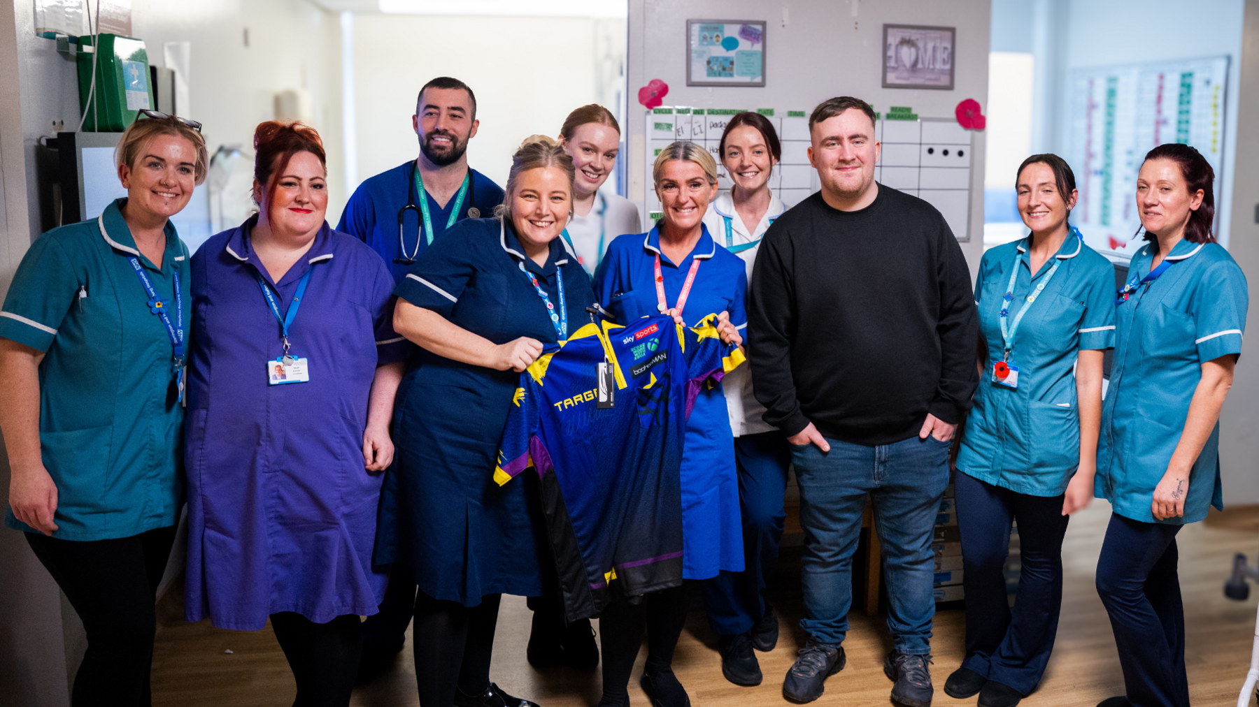 A group of healthcare staff in blue uniforms stand together in a hospital corridor posing with Luke Littler, who is in the middle. Medical charts and signage are visible on the walls behind them. Two members of staff hold a shirt presented by Littler.