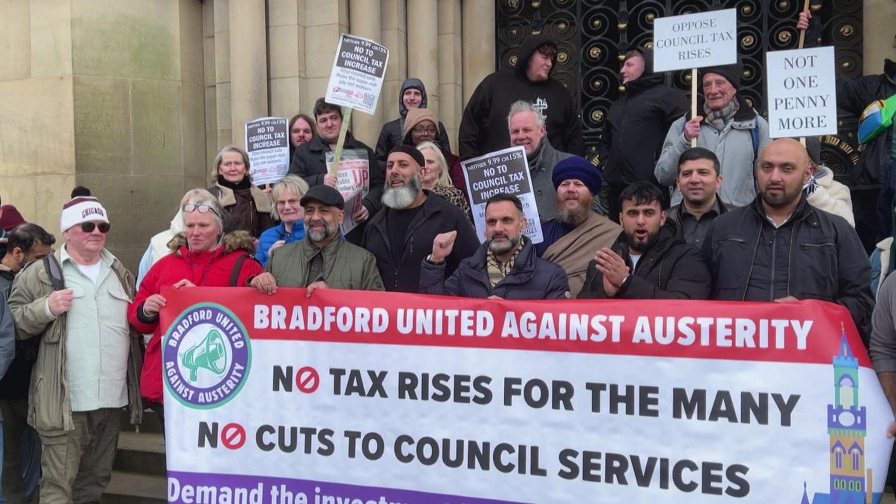 A group of people of mixed backgrounds and ages gathered on the steps of a town hall building, many of them holding banners and placards objecting to council tax rises.