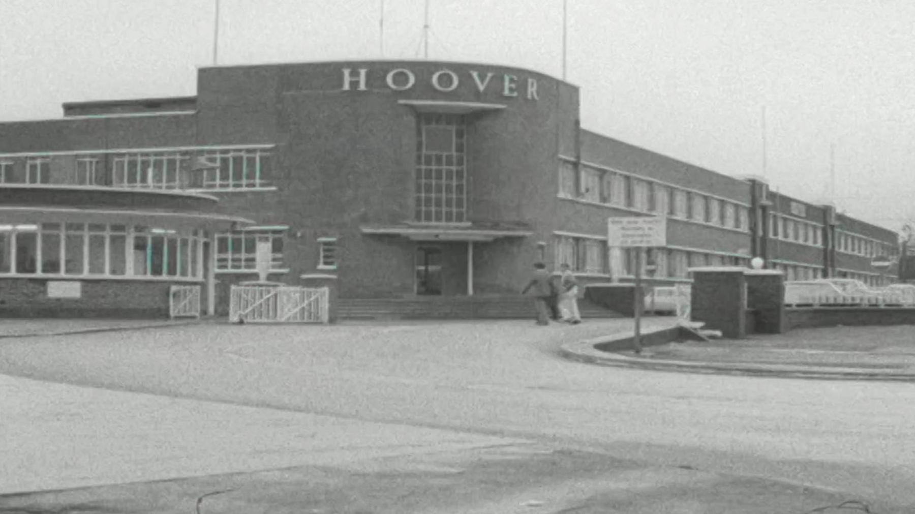 A general view of the Hoover factory in black and white. The entrance building is rounded in an art deco style with steps leading up to it. Cars can be seen in the courtyard as two people walk past on the street in front of it. 