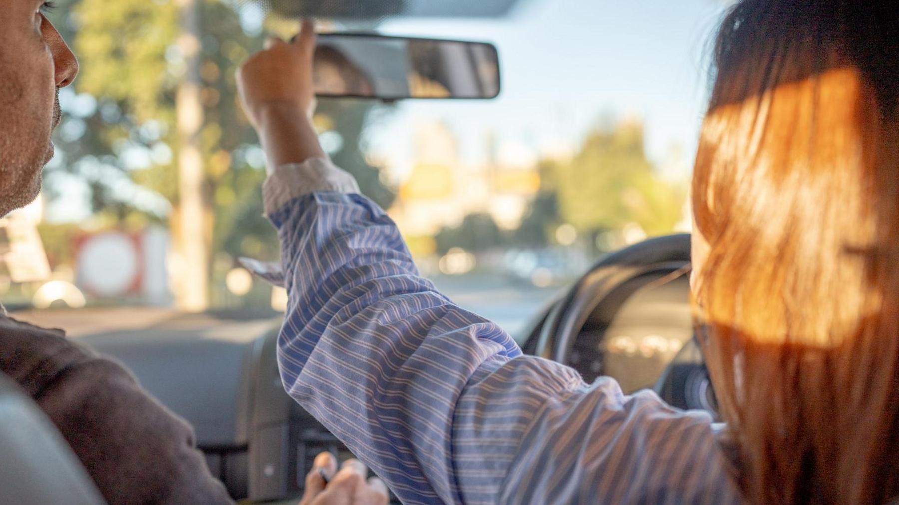 A woman sitting in the driver's seat of a car, adjusting her rear-view mirror with a male driving instructor sat next to her.