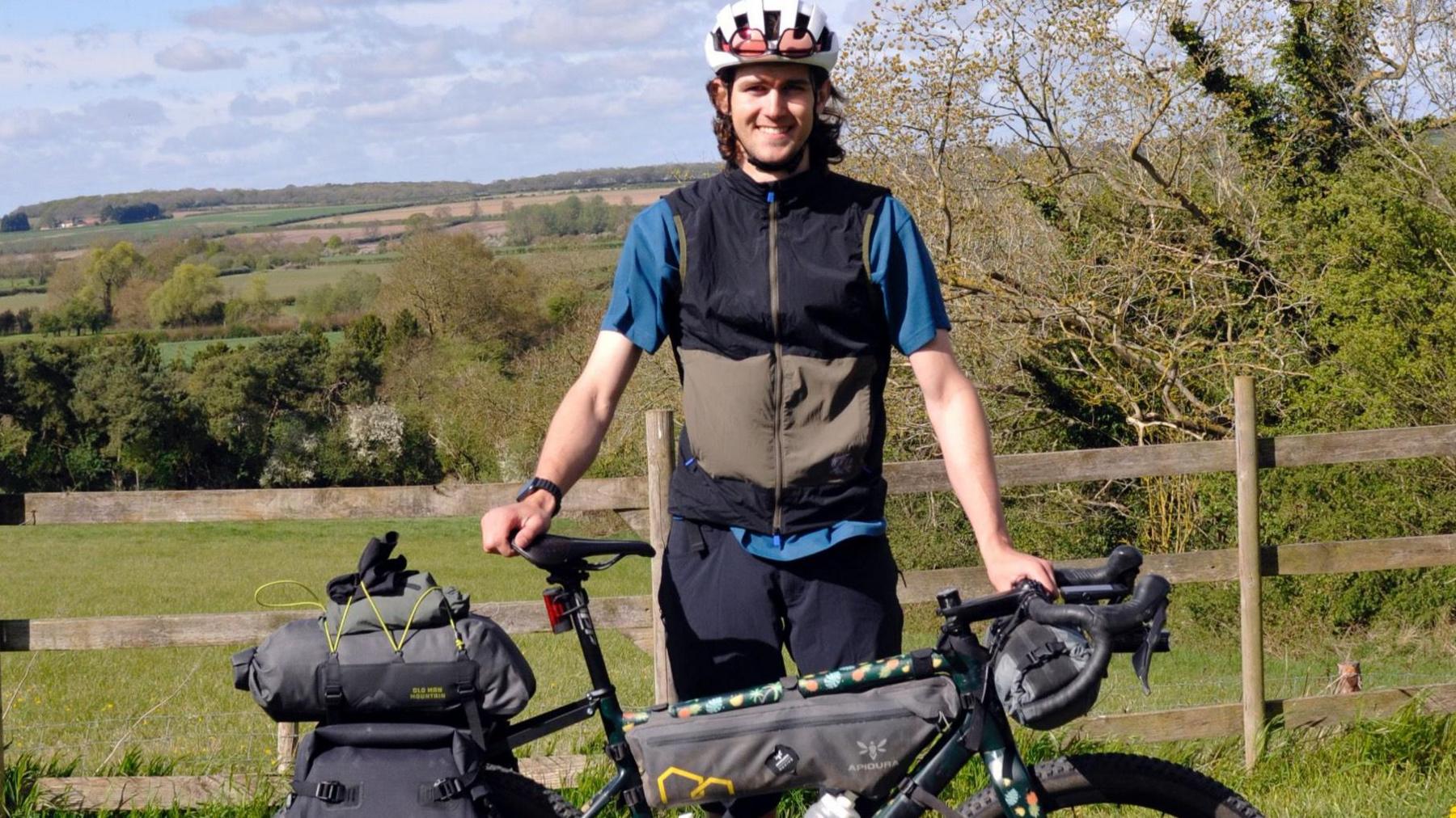 Finlay Tusting has brown, curly hair and is smiling directly at the camera while holding on to his bike packed with equipment. He is standing in front of an English country scene, with gentle hills in the background.