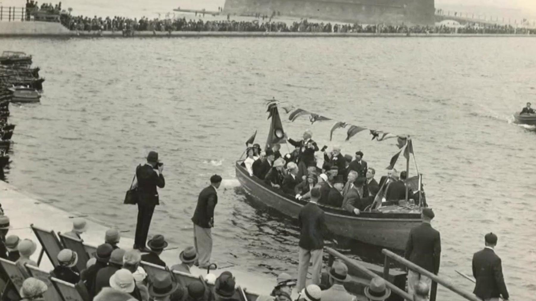 A boat carrying a number of passengers is sailing across the water at the New Brighton Marine Lake in this black and white photograph. A number of spectators watch - some in deckchairs while others are standing.