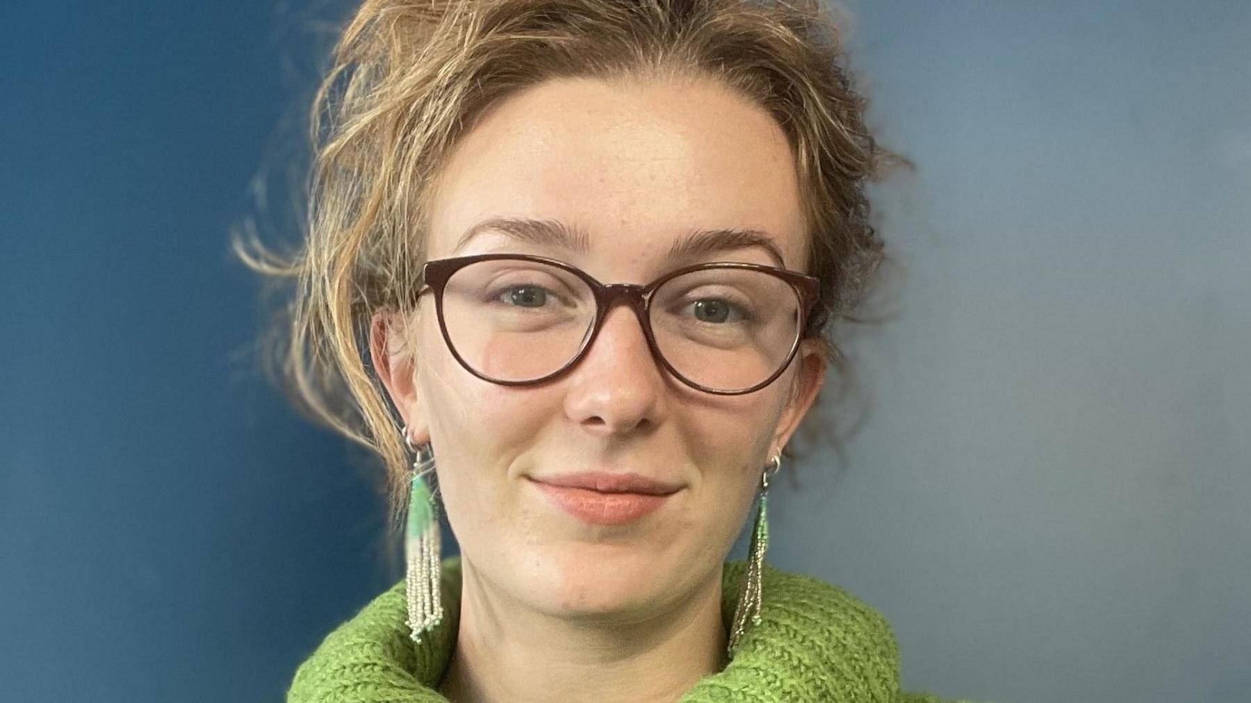 Sarah Peart is wearing dark-rimmed glasses and a green, knitted roll-neck jumper, and dangly earrings. She has blonde hair which is tied up and is stood against a blue wall.