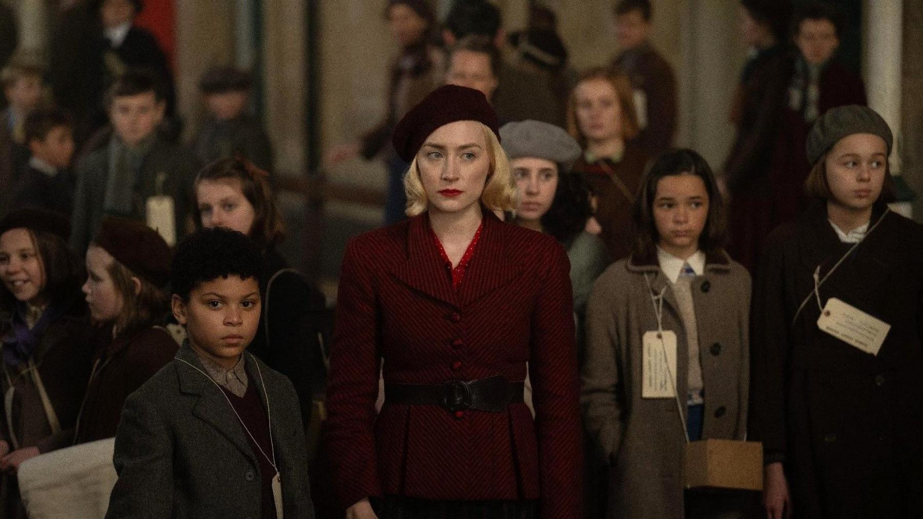 A still from the film Blitz, with a woman and a boy dressed in 1940s clothing. They are surrounded by children playing evacuees. The woman is wearing a red coat with a black belt around her waist and with a matching red beret. The children have what appears name tags around their neck.