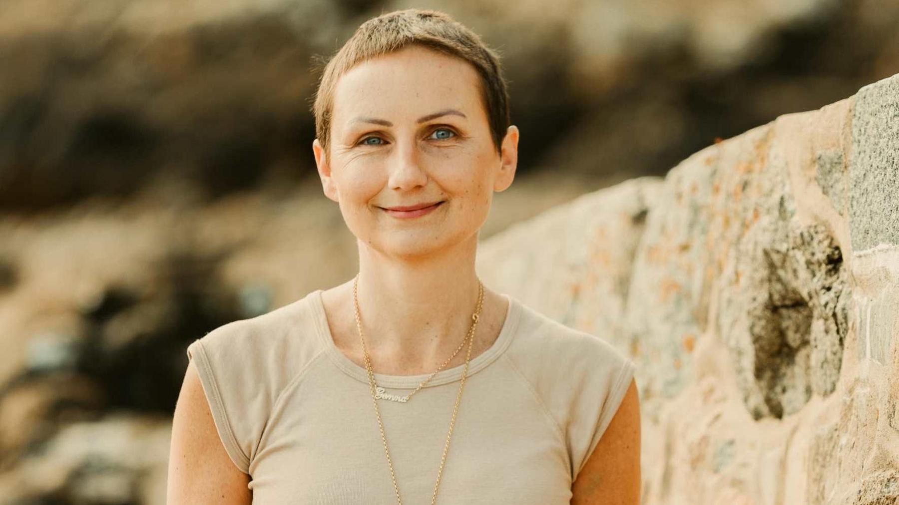Gemma Ainger is pictured smiling. She has short brown hair and blue eyes and is smiling at the camera. She is wearing a beige T-shirt and two gold necklaces, one of which says Gemma in gold cursive writing. She appears to be standing in front of some rocks, perhaps on a beach.