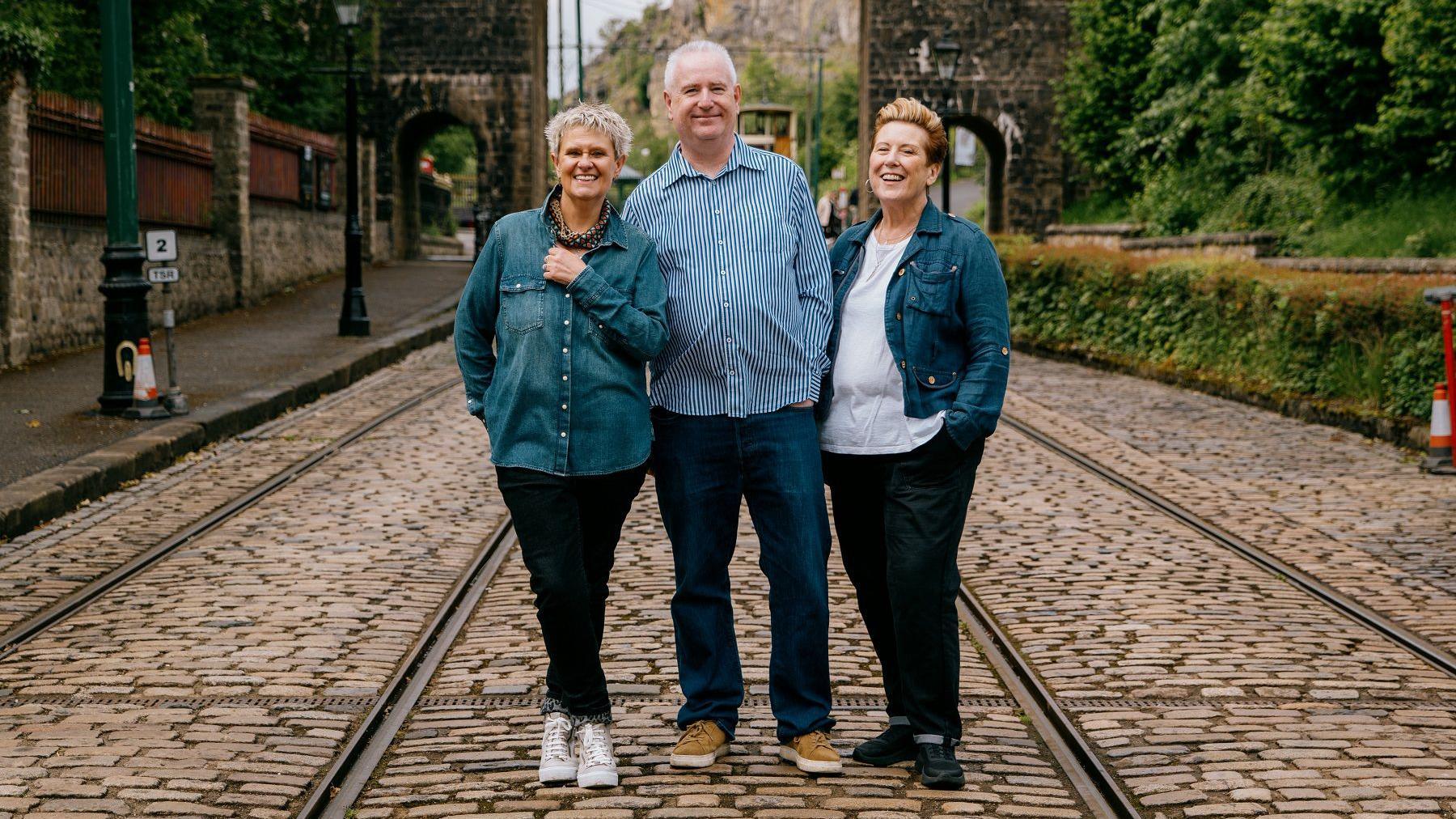 Two women wearing denim shirts and black trousers, and a man wearing jeans and a blue and white stripy shirt, standing on a train track and smiling. There's vegetation and a bridge behind them.