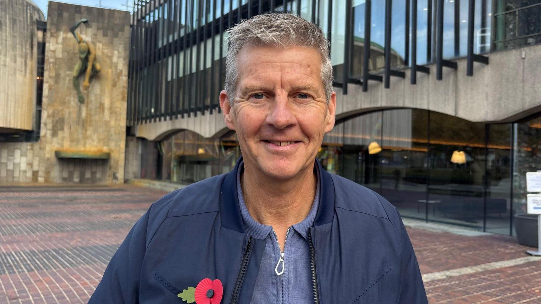 Steve Cram is wearing a blue jacket with a poppy on the lapel, and is standing in front of the Newcastle Civic Centre smiling at the camera.