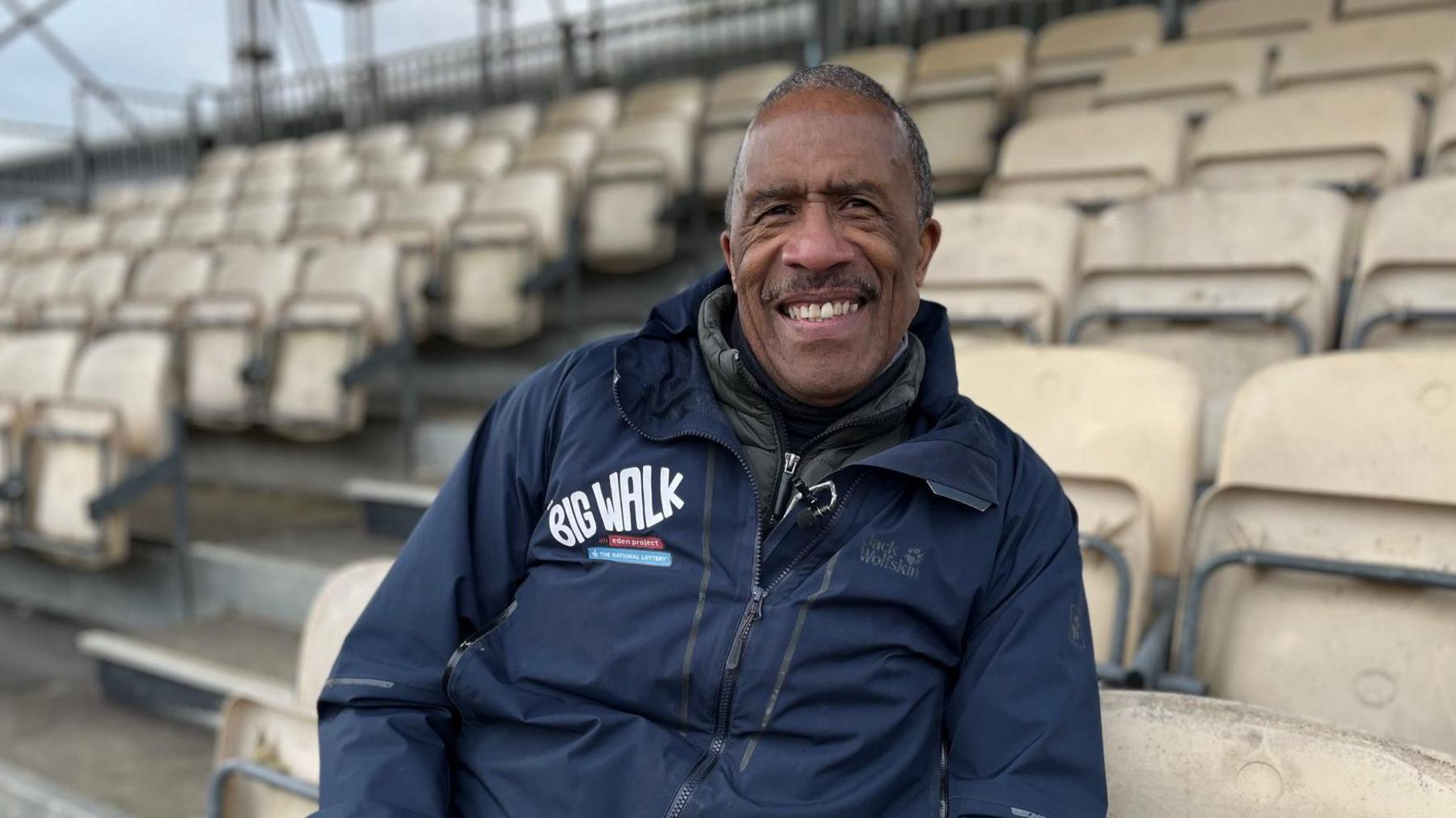 An elderly man sits on white chairs in a football stadium and smiles at the camera.  