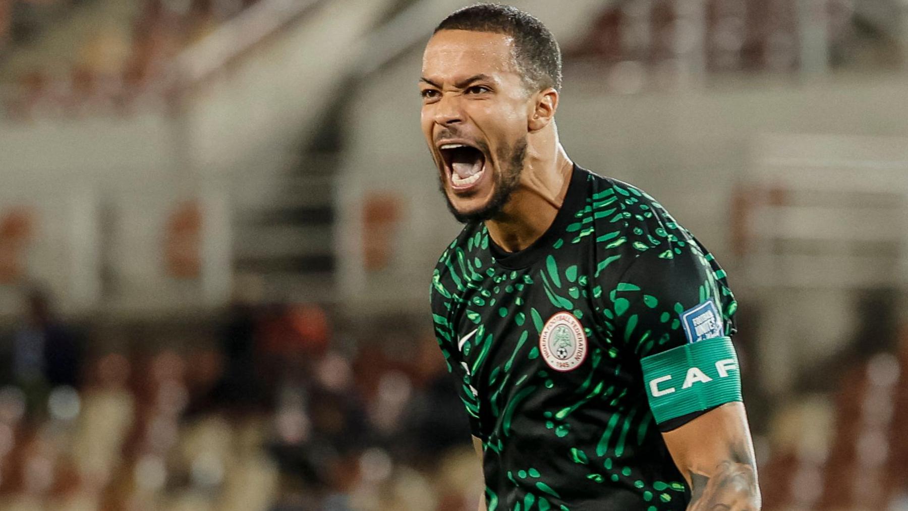 William Troost-Ekong screams in delight as he celebrates scoring a penalty for Nigeria in a stadium where a largely empty stand is blurred in the background