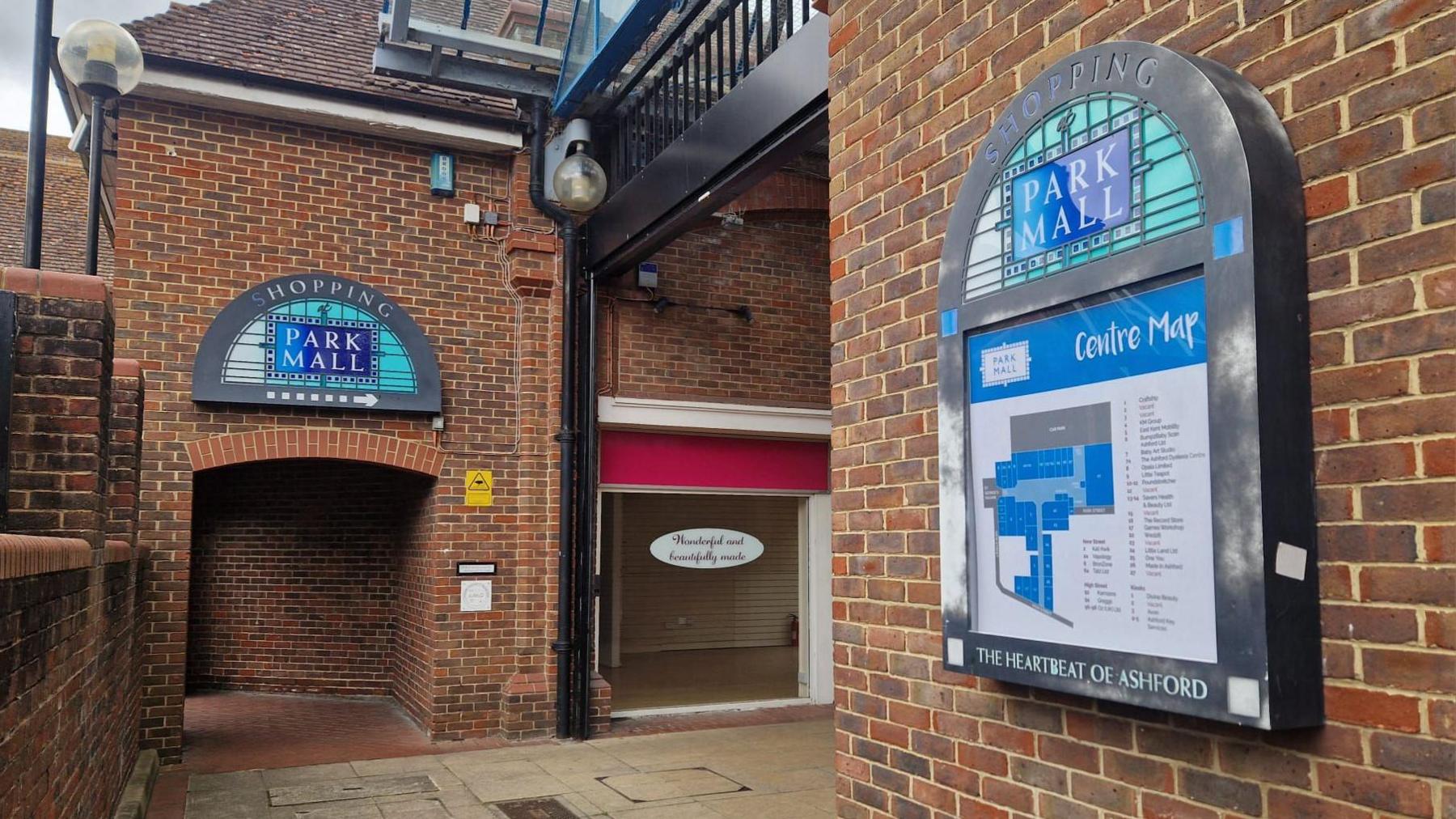 An entrance to an outdoor shopping centre. There is a large map on one wall showing the plan of the centre and in the back ground, an empty shop, with an old sign in the glass window.