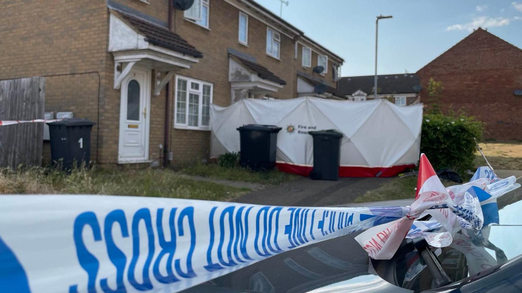 Blue and white police tape cordoning off the entrance to the house, which is mid-terrace and made of cream bricks. There is a white tent surrounding the entrance.
