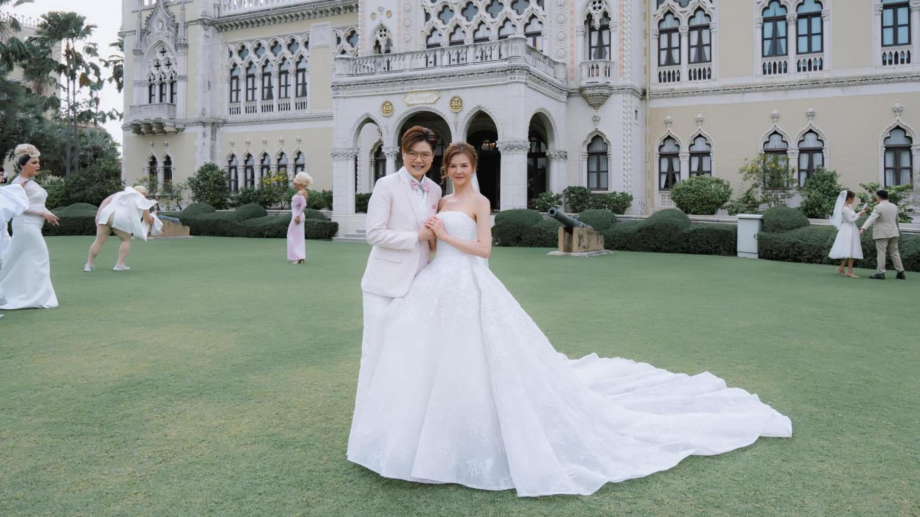 Rungtiwa in a white wedding gown and Phanlavee in a white suit with a pink corsage. Both are smiling in a lawn in front of a stately white building.