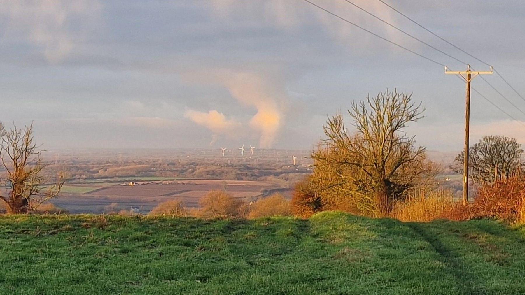 A distant view of countryside from the Cotswolds in Gloucestershire, under winter sunshine. On the horizon is a distinct plume of white cloud rising up high into the sky