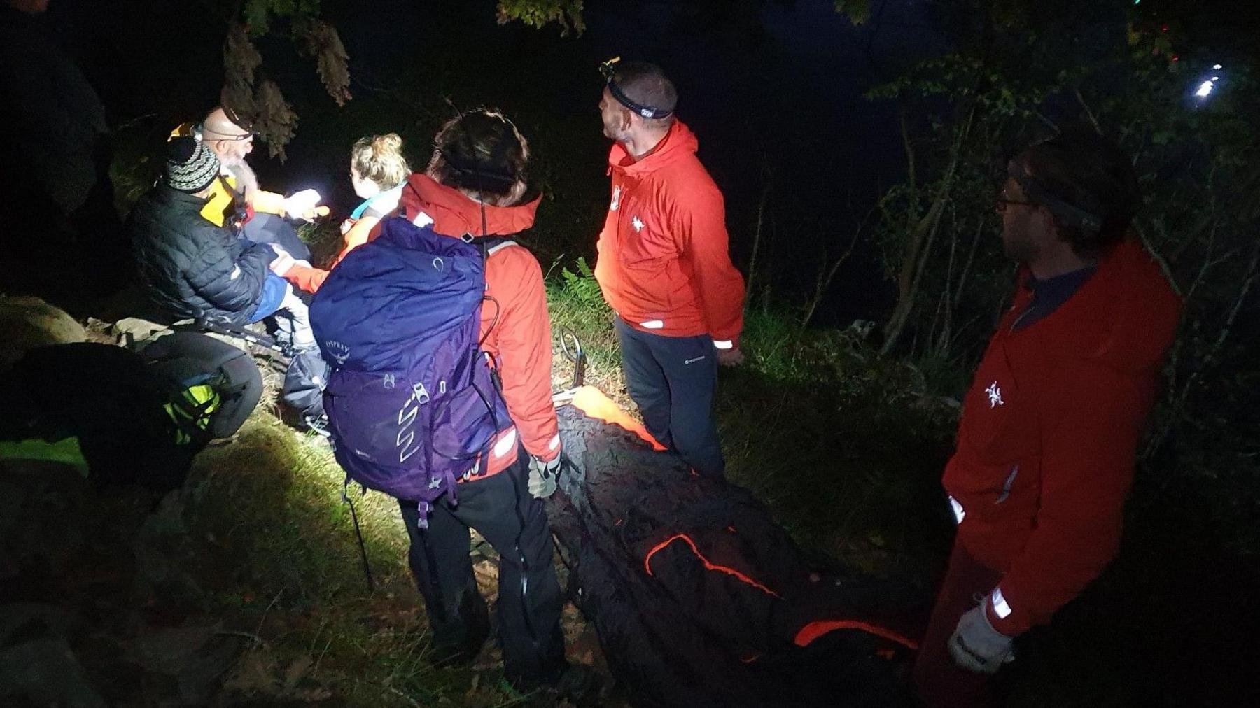 A nighttime rescue scene shows several people, some in red jackets, assisting an individual on a stretcher near a body of water with flashlights illuminating the area.