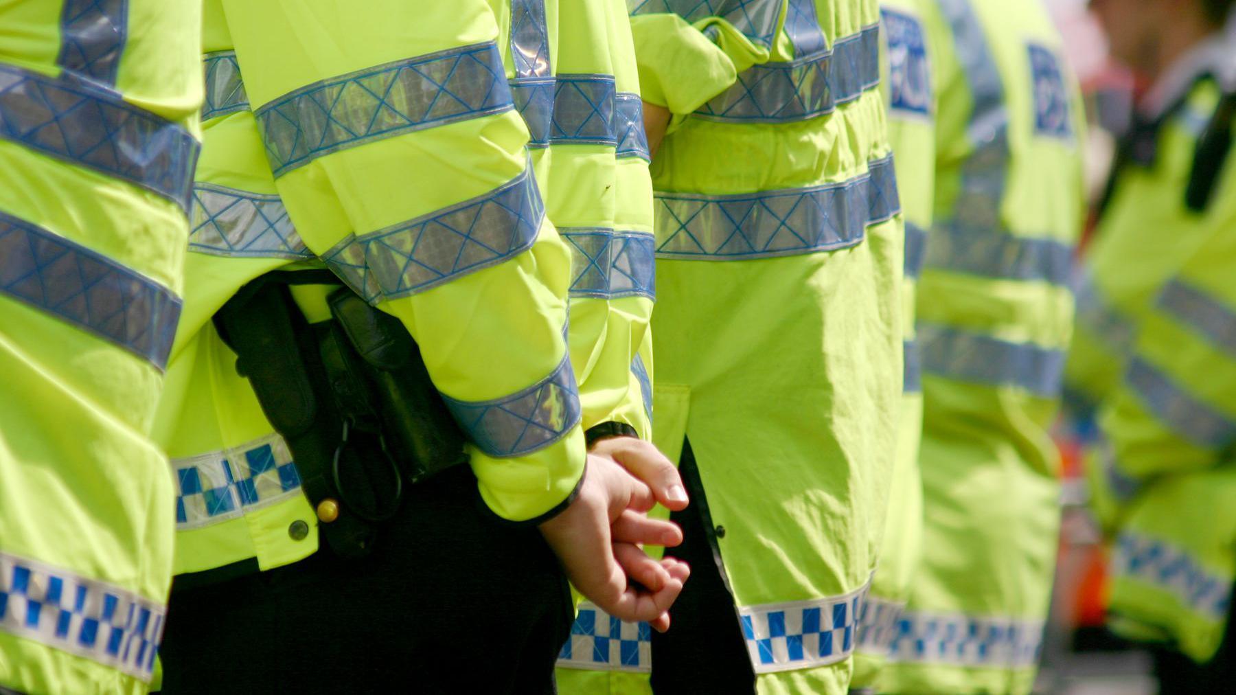 A row of police officers wearing yellow high vis long-sleeved jackets. One with hands held behind their back.