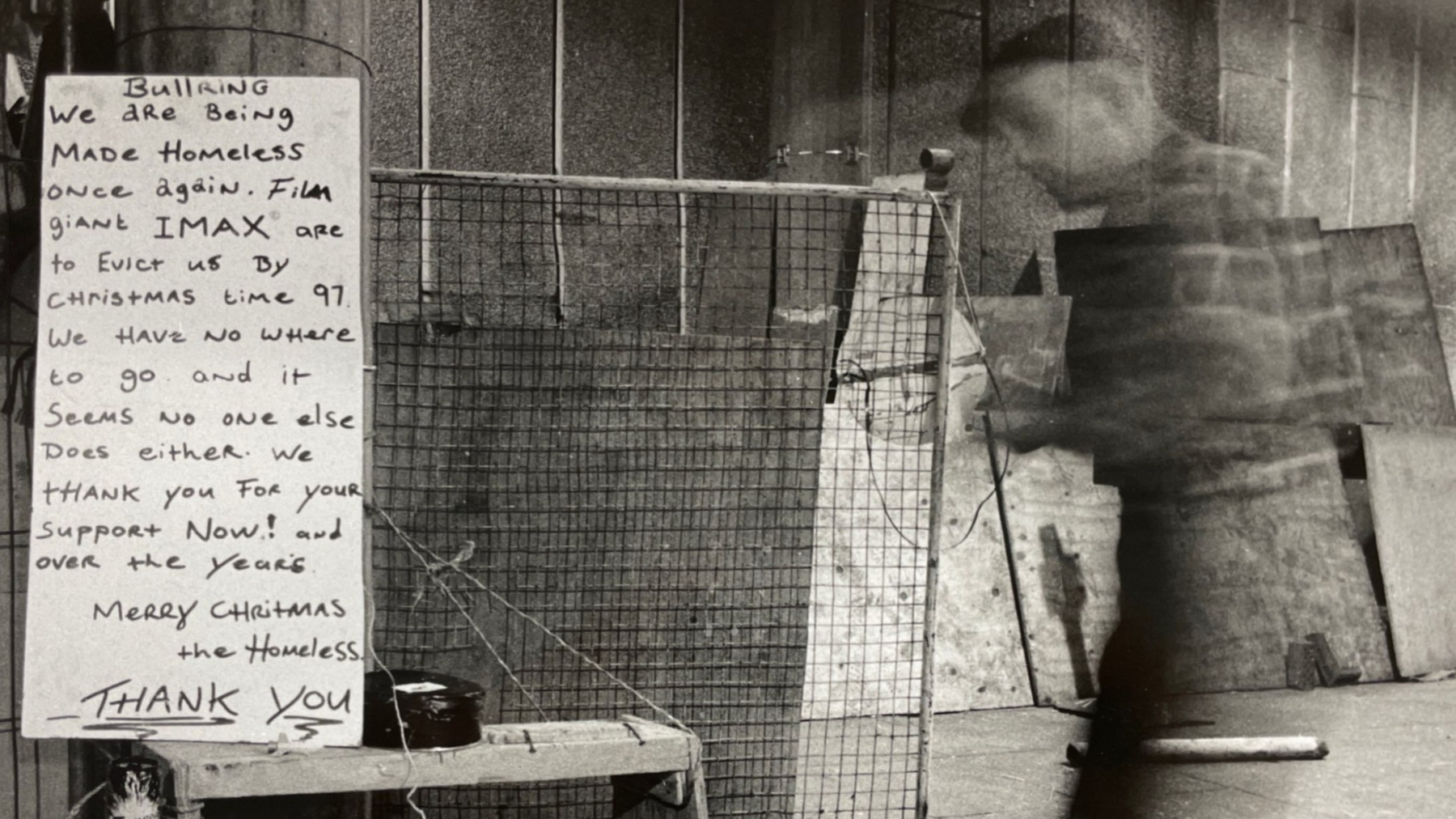 A black and white stylised photo showing a blurred man walking past a wire fence and handwritten sign in a tunnel. The sign says 'Bullring: we are being made homeless once again. Film giant Imax are to evict us by Christmastime '97. We have nowhere to go and it seems no-one else does either. We thank you for your support now and over the years. Merry Christmas, the homeless. Thank you'