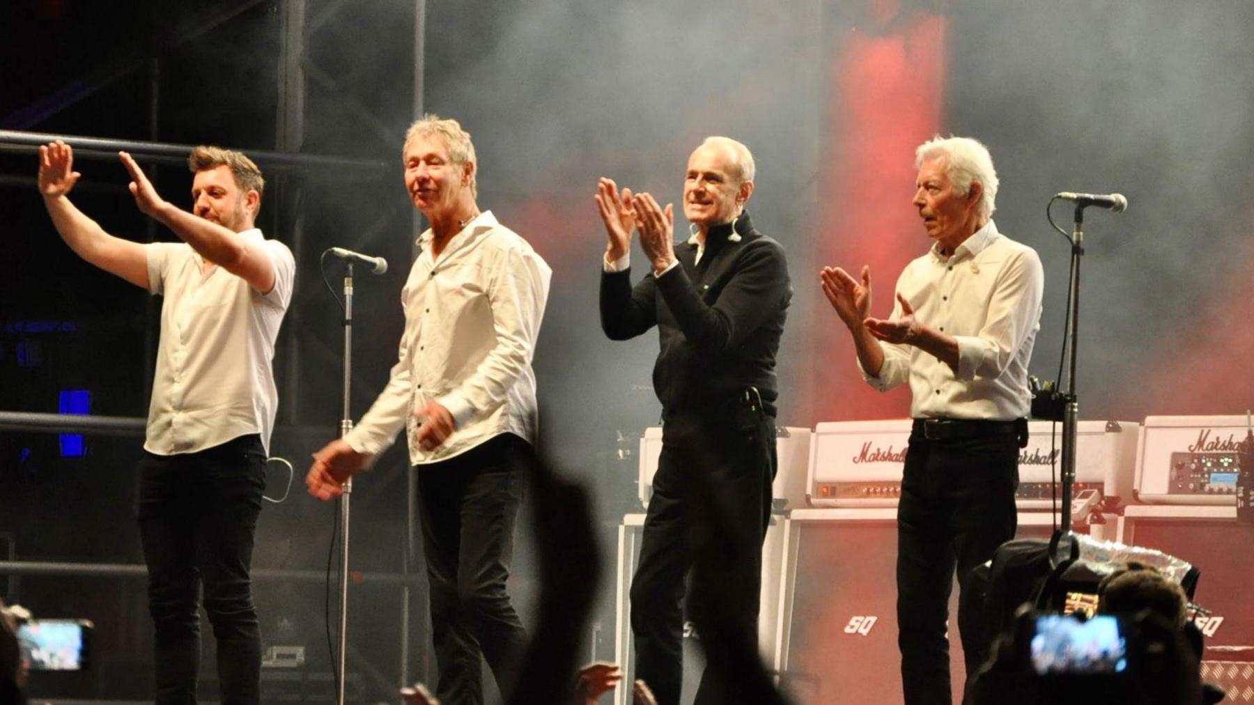 A picture of rock band Status Quo on stage at Taunton's Vivary Park.  They are clapping and thanking the crowd.