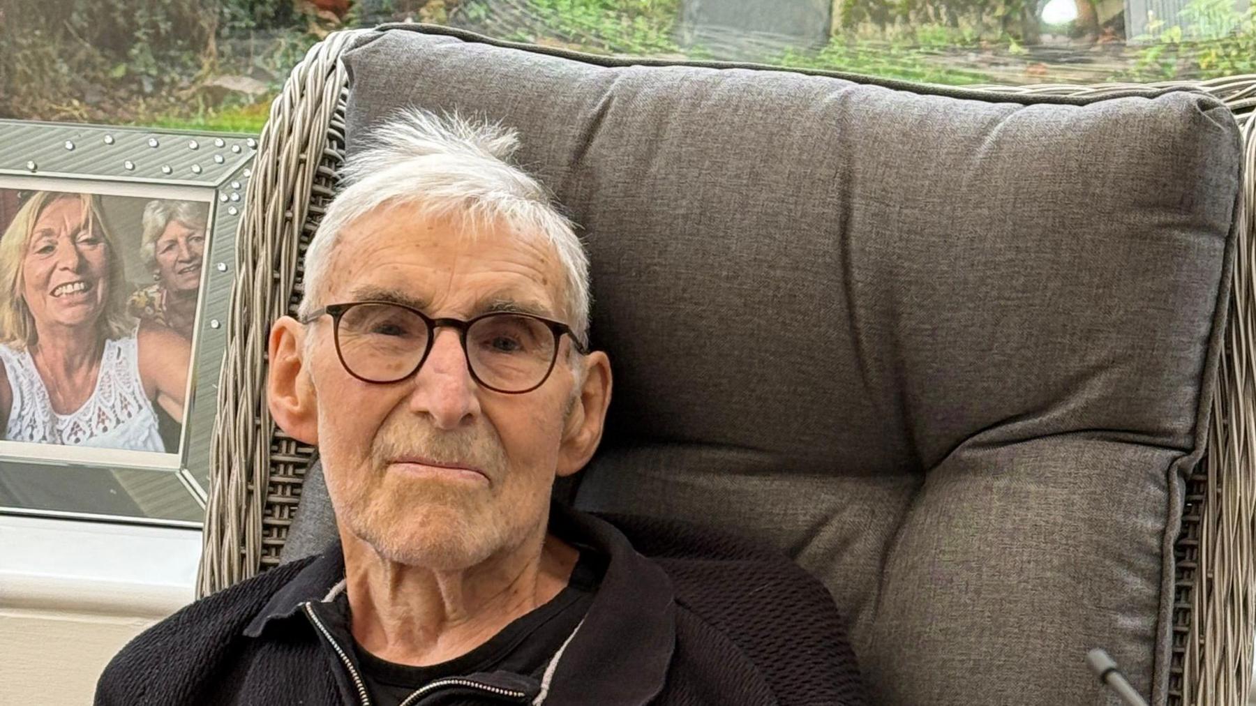 Ray Day smiling towards the camera and sitting in a grey chair. He is wearing a black jumper. In the background is windows and photographs along the window ledge.