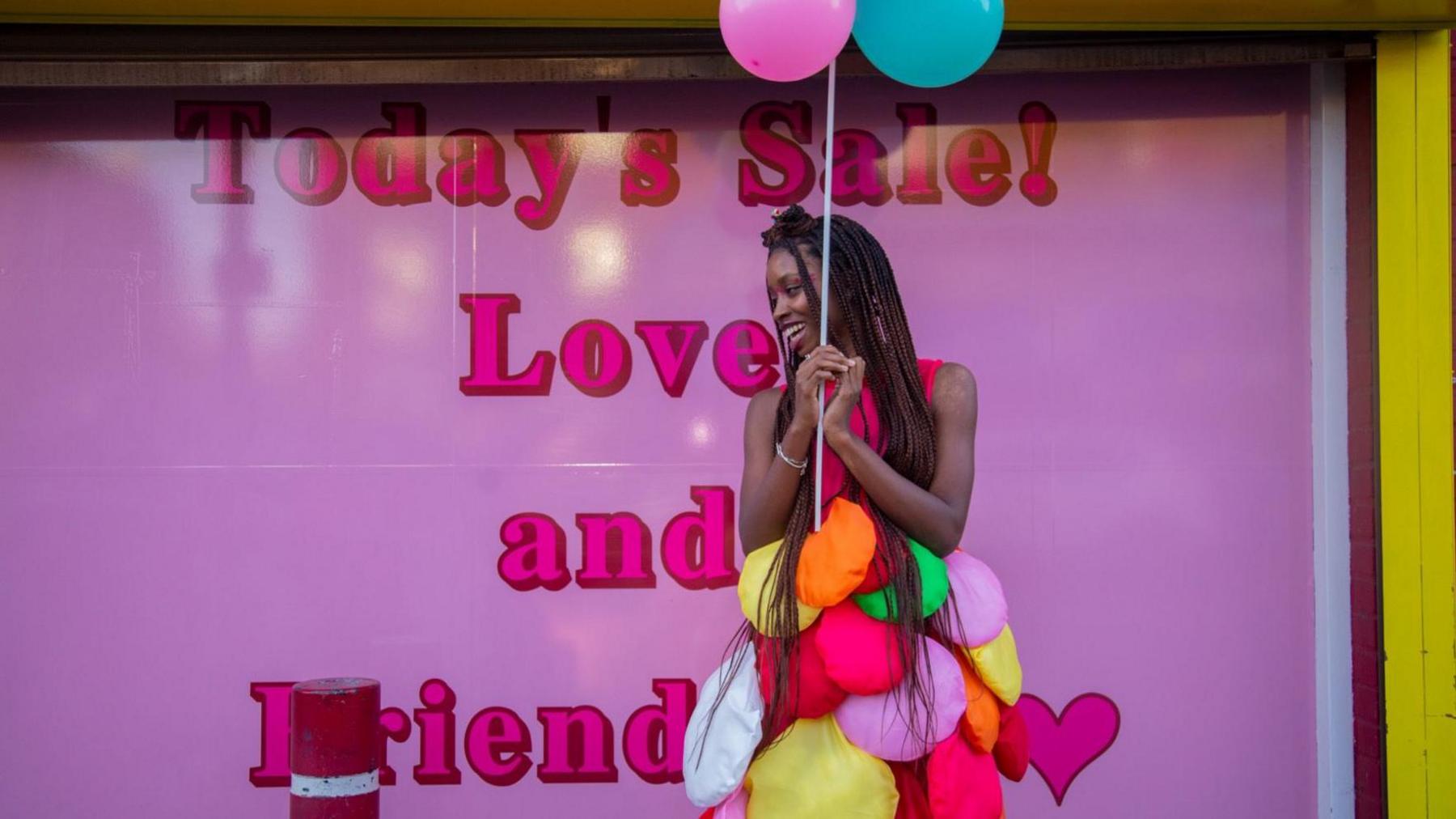 A woman stood in front of a pink poster wearing a colourful outfit.