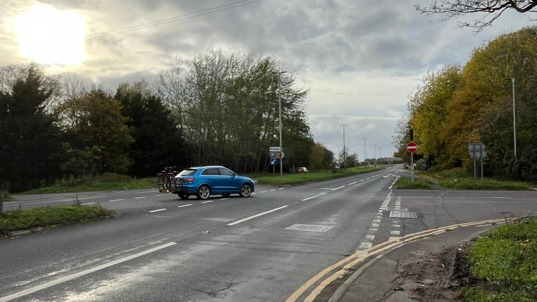A view down a dual carriageway on a grey day: there is a junction on the right hand side, with a car in a section in the middle of the road, about to turn right into it.