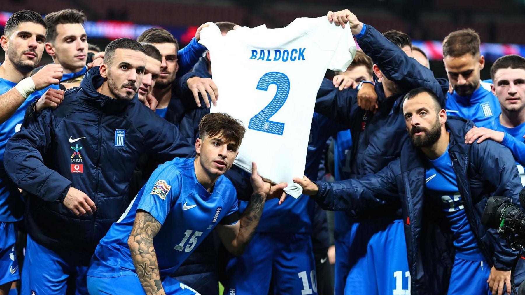 Greece players hold up a shirt bearing George Baldock's name and number two after their victory against England