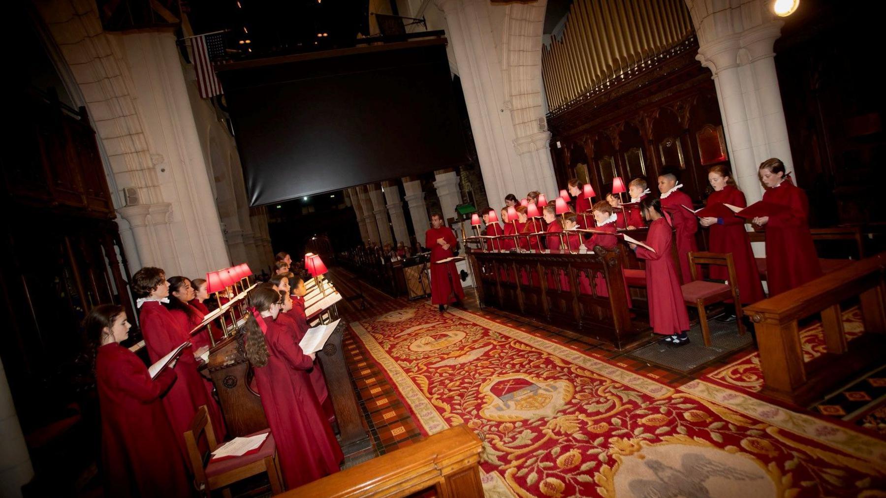 A group of children choristers are dressed in red gowns as they sing. They are standing in wooden pews and holding red song books. They are singing within a church which has white pillars and wooden details. 