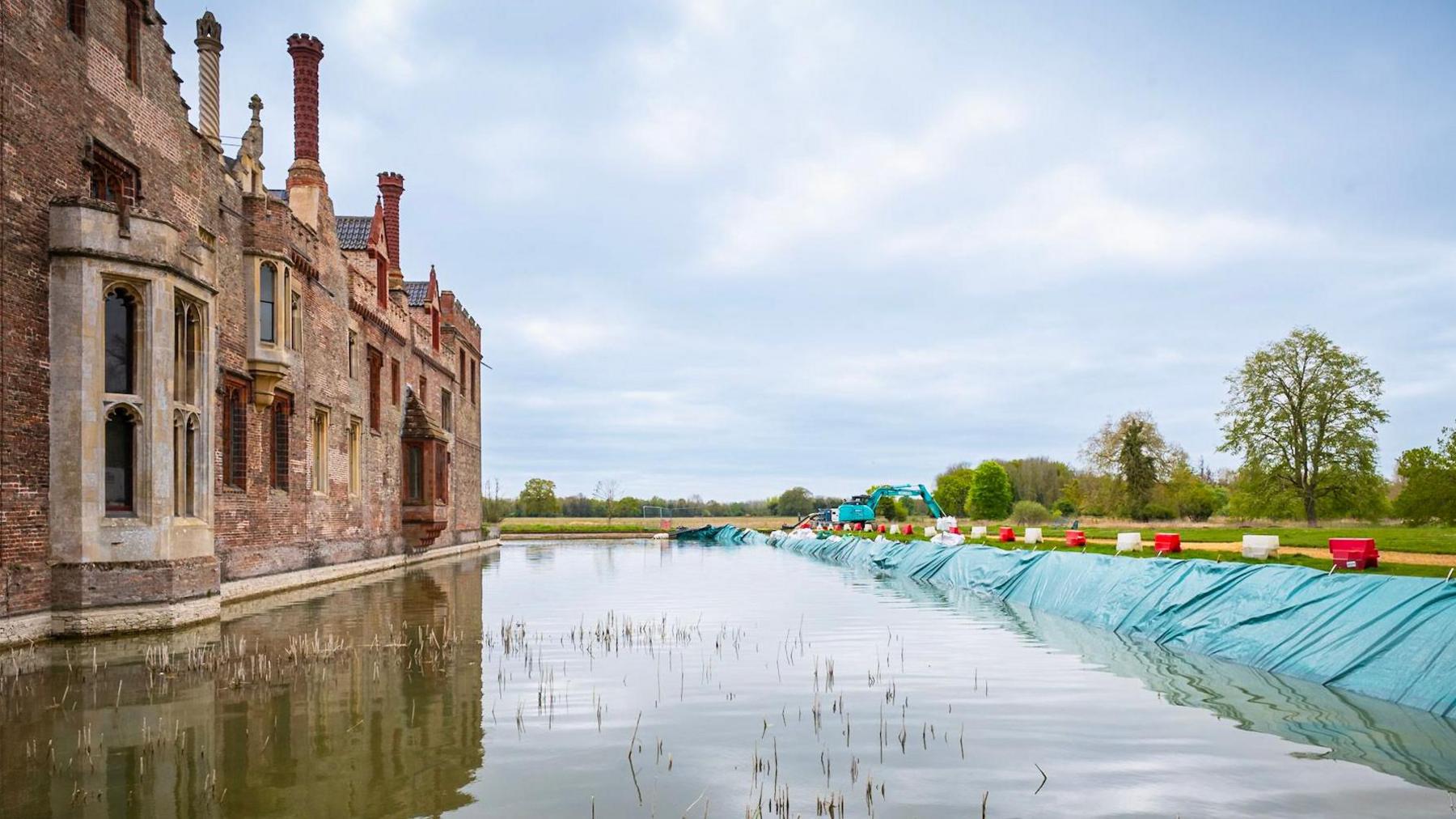 Red brick Oxburgh Hall is on the left hand side of the photograph. The moat is featured in the middle and on the right is green, plastic sheeting running the length of the moat and holding the water in place.