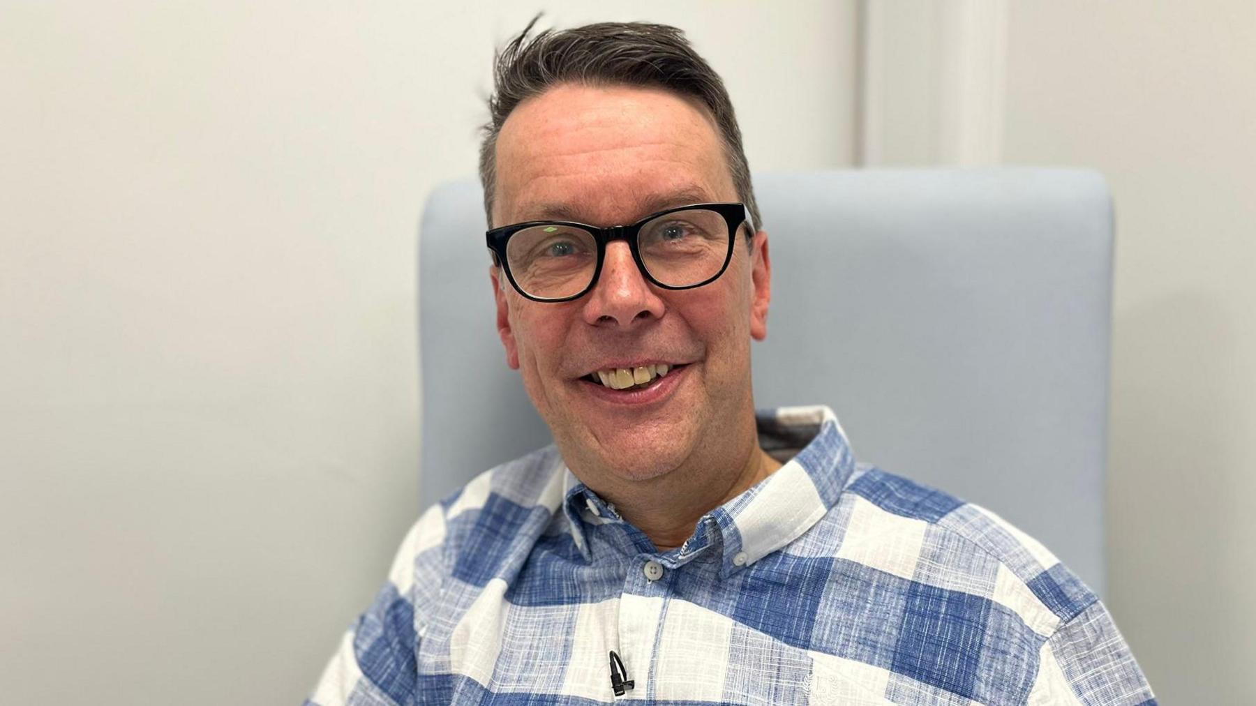 Paul Silvester smiling in a chair wearing a blue and white checked linen shirt and black thick-rimmed glasses